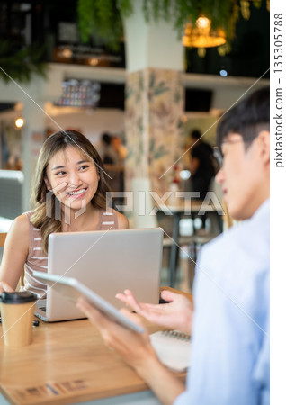 Asian woman looking at a man holding tablet talking as sits with laptop at table in cafe or canteen Asian woman looking at a man holding tablet talking as sits with laptop at table in cafe or canteen 135305788