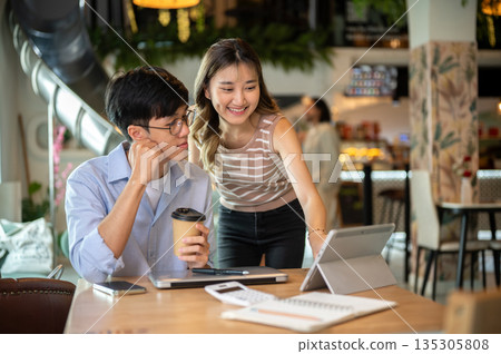 Asian woman standing and bending over wooden table looking at a man's tablet in cafe or canteen. 135305808