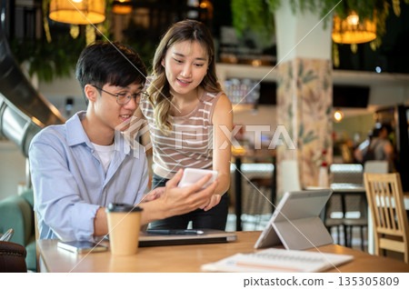 Asian woman standing and bending over wooden table looking at a man's calculator in cafe or canteen. Asian woman standing and bending over wooden table looking at a man's calculator in cafe or canteen. 135305809