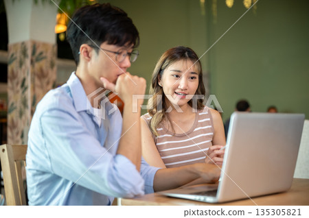 Glasses asian man and woman thinking as looking at laptop sitting at wooden table in cafe or canteen Glasses asian man and woman thinking as looking at laptop sitting at wooden table in cafe or canteen 135305821