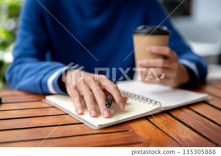 Close up of hand with pen and other holding coffee over notebook at wood table in cafe or canteen. Close up of hand with pen and other holding coffee over notebook at wood table in cafe or canteen. 135305888