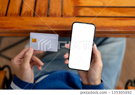 Close up of a man looking at phone holding credit card in hand at wooden table in cafe or canteen. Close up of a man looking at phone holding credit card in hand at wooden table in cafe or canteen. 135305892