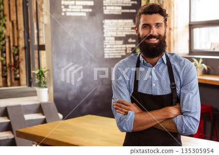 Adult male barista wearing black apron smiling in coffee shop showing chalkboard menu, copy space Adult male barista wearing black apron smiling in coffee shop showing chalkboard menu, copy space 135305954