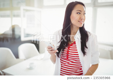 Asian female standing near white table with coffee cup holding smartphone in office, copy space 135305966