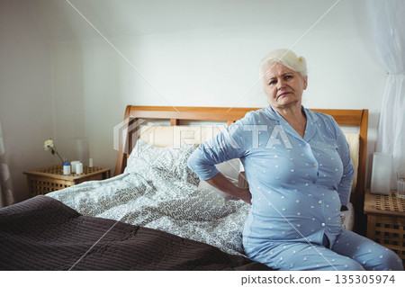 Senior woman wearing pajamas sitting at bedside holding lower back with pill bottles, copy space 135305974