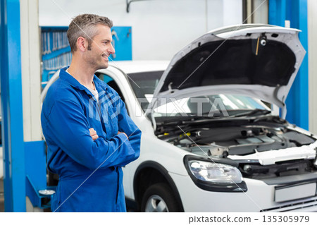 Male mechanic standing in car repair garage folding arms and smiling beside open white car hood Male mechanic standing in car repair garage folding arms and smiling beside open white car hood 135305979