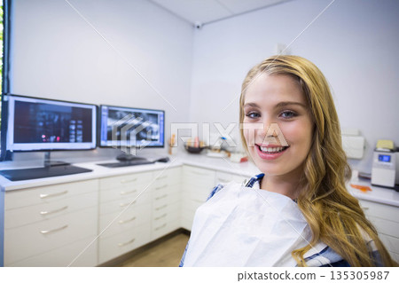 Female patient sitting in dental clinic wearing bib, viewing X-rays on dual monitors, copy space 135305987