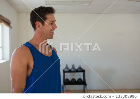 Man standing in gym wearing blue tank top beside kettlebell rack and window blinds, copy space Man standing in gym wearing blue tank top beside kettlebell rack and window blinds, copy space 135305996
