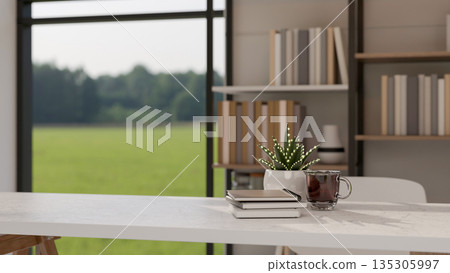 Glass of coffee and book aside pot plant on white table in a bright living room with greenery view. 135305997