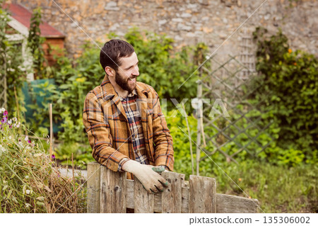 Male gardener leaning on fence in backyard among flowering plants wearing plaid shirt and gloves 135306002