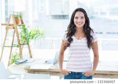 Woman standing by desk in office using laptop amid notebooks, papers and plant stand, copy space 135306007