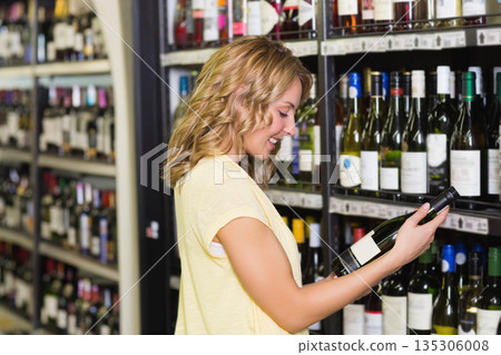Woman holding wine bottle at chest height tilting label in liquor store aisle with price tags Woman holding wine bottle at chest height tilting label in liquor store aisle with price tags 135306008