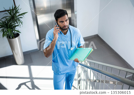 Asian male worker with stethoscope climbing hospital stairwell holding smartphone, colored folders Asian male worker with stethoscope climbing hospital stairwell holding smartphone, colored folders 135306015