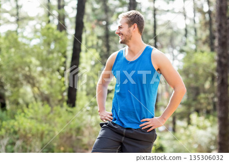 Man in sportswear standing hands on hips, smiling and looking left in forest clearing, copy space 135306032
