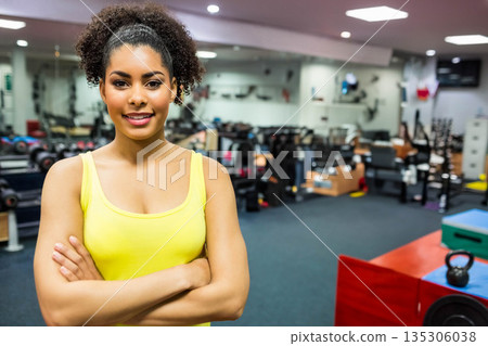 Woman crossing arms and smiling while standing in gym with dumbbells, yellow tank top, copy space 135306038