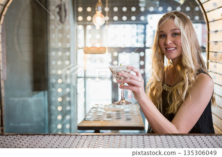 Blonde teenage girl sitting under Edison bulbs at bar holding cocktail glass with herb, copy space Blonde teenage girl sitting under Edison bulbs at bar holding cocktail glass with herb, copy space 135306049