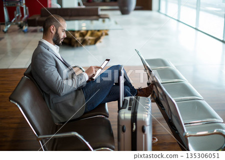 White male businessman sitting at airport lounge using tablet beside suitcase, copy space White male businessman sitting at airport lounge using tablet beside suitcase, copy space 135306051