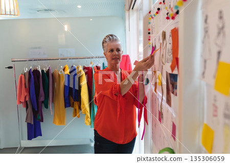 Senior female fashion designer pinning swatch to board in bright design studio with garment rack 135306059