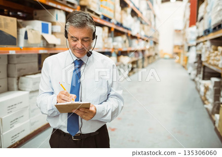 Male warehouse supervisor holding clipboard and pencil, wearing headset checking inventory in aisle 135306060
