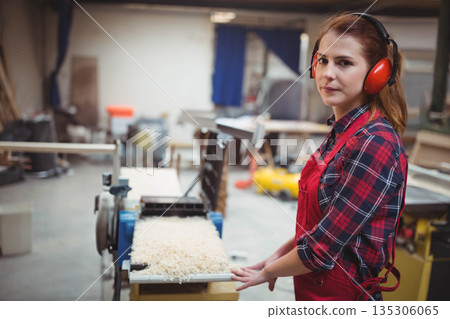 Female carpenter with red apron and earmuffs using planer in workshop making shavings, copy space 135306065