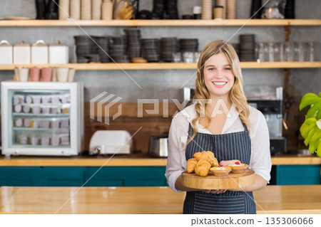 Female barista serving pastries on wooden tray behind café counter and display case, copy space 135306066