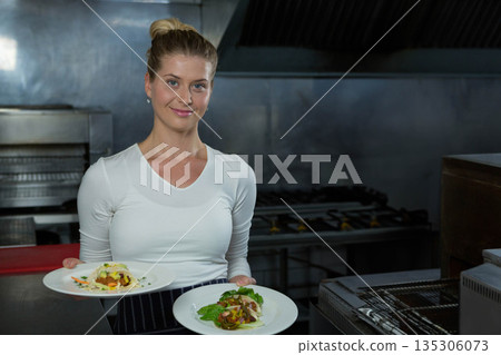 Female chef wearing striped apron and white shirt holding two salad plates in commercial kitchen 135306073
