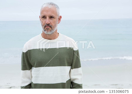 Senior man standing on sandy shoreline at beach wearing olive green striped crewneck sweater 135306075