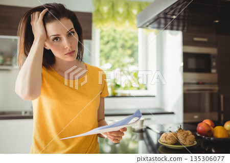 Woman holding documents and scratching head at kitchen countertop near pastries and fruit bowl Woman holding documents and scratching head at kitchen countertop near pastries and fruit bowl 135306077