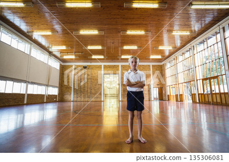 School-aged boy standing on wooden gym floor wearing white polo with navy shorts under tall windows 135306081
