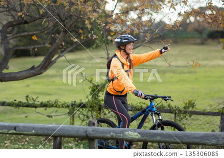 Woman riding mountain bicycle and pointing forward by fence in park wearing helmet and backpack Woman riding mountain bicycle and pointing forward by fence in park wearing helmet and backpack 135306085