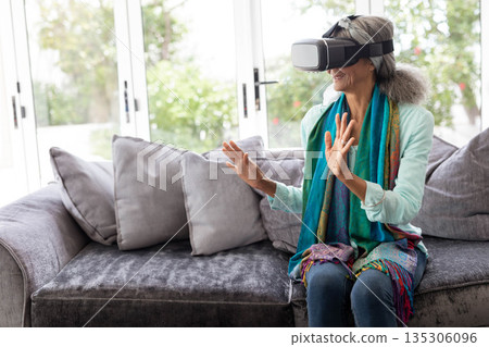 Senior woman wearing VR headset and colorful scarf sitting on sofa by glass doors revealing garden 135306096