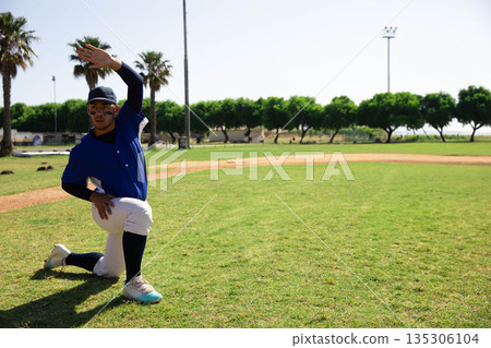 Teenage boy stretching right arm while kneeling on baseball field near dirt infield, copy space Teenage boy stretching right arm while kneeling on baseball field near dirt infield, copy space 135306104