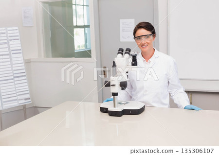 Female scientist wearing lab coat and gloves examining sample under stereo microscope on lab bench 135306107