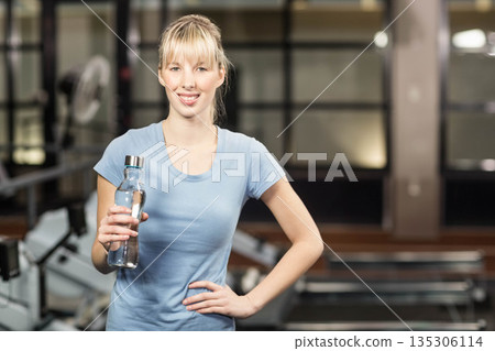 Woman holding water bottle and resting on hip inside gym near rowing machines and treadmills Woman holding water bottle and resting on hip inside gym near rowing machines and treadmills 135306114