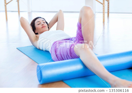 Woman performing crunches using foam roller on blue yoga mat in fitness studio with barre supports 135306116