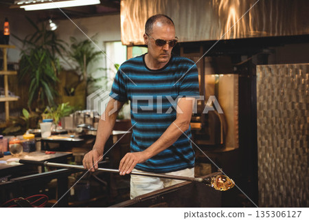 Middle-aged man standing at glassblowing bench gripping blowpipe with molten glass at furnace Middle-aged man standing at glassblowing bench gripping blowpipe with molten glass at furnace 135306127