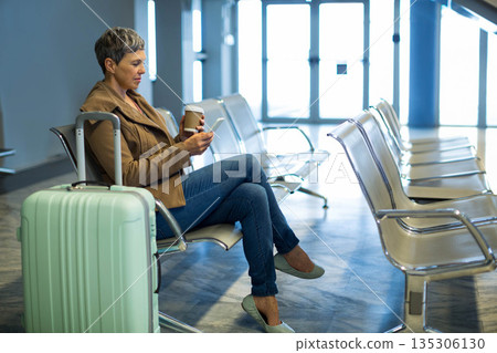 Senior woman with rolling suitcase sitting on airport seats holding coffee cup and using smartphone Senior woman with rolling suitcase sitting on airport seats holding coffee cup and using smartphone 135306130