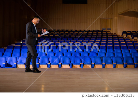 Asian mature man presenting on stage floor beside blue seats in hall holding papers, copy space Asian mature man presenting on stage floor beside blue seats in hall holding papers, copy space 135306133