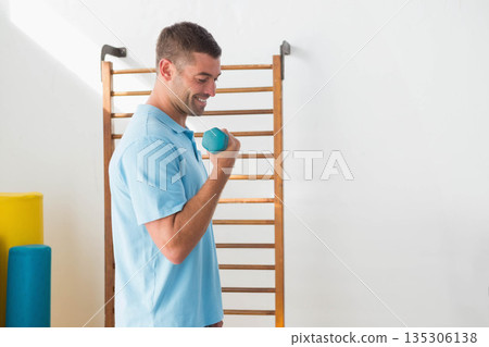 Man lifting hex dumbbell by stall bars with foam rollers in rehab room wearing polo shirt Man lifting hex dumbbell by stall bars with foam rollers in rehab room wearing polo shirt 135306138