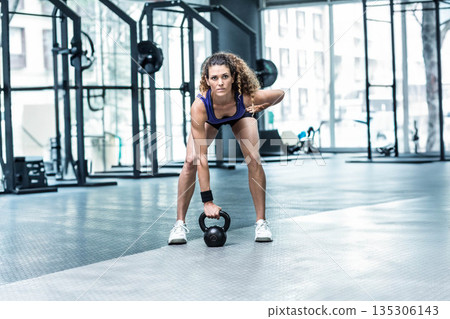 Female fitness enthusiast wearing blue tank top lifting black kettlebell while bending over in gym Female fitness enthusiast wearing blue tank top lifting black kettlebell while bending over in gym 135306143