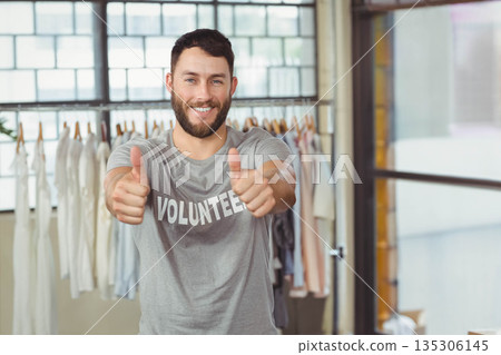 Mid adult man wearing volunteer shirt giving thumbs up by rack with shirts in donation area 135306145