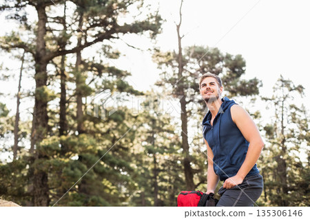 Man kneeling on rocky outcrop in pine forest wearing navy hoodie gripping red backpack, copy space Man kneeling on rocky outcrop in pine forest wearing navy hoodie gripping red backpack, copy space 135306146