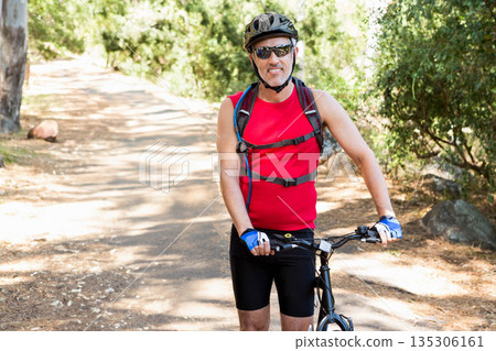 Male cyclist standing on sunlit forest trail wearing helmet and hydration pack with bicycle 135306161