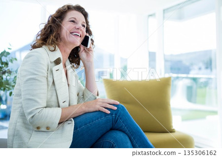 Middle-aged woman wearing blazer on sofa in living room holding phone beside cushion, copy space 135306162