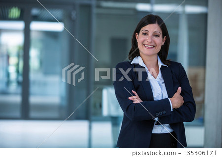 Mature businesswoman wearing suit posing folding arms and smiling in office lobby glass doors Mature businesswoman wearing suit posing folding arms and smiling in office lobby glass doors 135306171