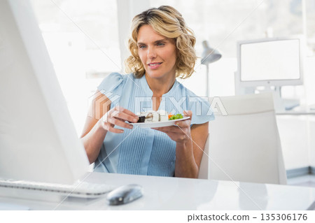 Woman holding sushi plate with chopsticks while using computer at modern office workspace Woman holding sushi plate with chopsticks while using computer at modern office workspace 135306176