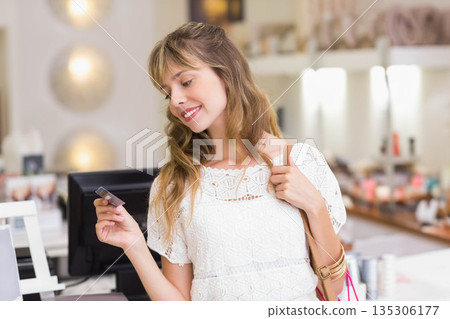 Woman standing at cosmetics counter holding credit card by card reader and shopping bags 135306177