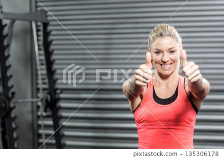 Athletic woman smiling and giving thumbs up in gym with barbell rack, weight plates and shutter 135306178