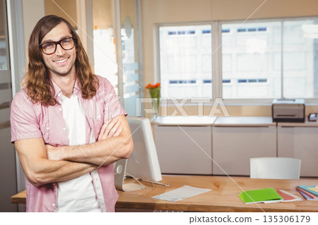 Mid adult man standing arms crossed at desk in office with computer monitor, tulips, copy space Mid adult man standing arms crossed at desk in office with computer monitor, tulips, copy space 135306179