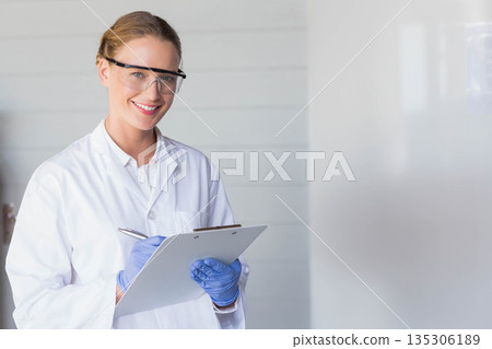 Female technician wearing lab coat goggles and gloves using clipboard with pen in lab, copy space Female technician wearing lab coat goggles and gloves using clipboard with pen in lab, copy space 135306189